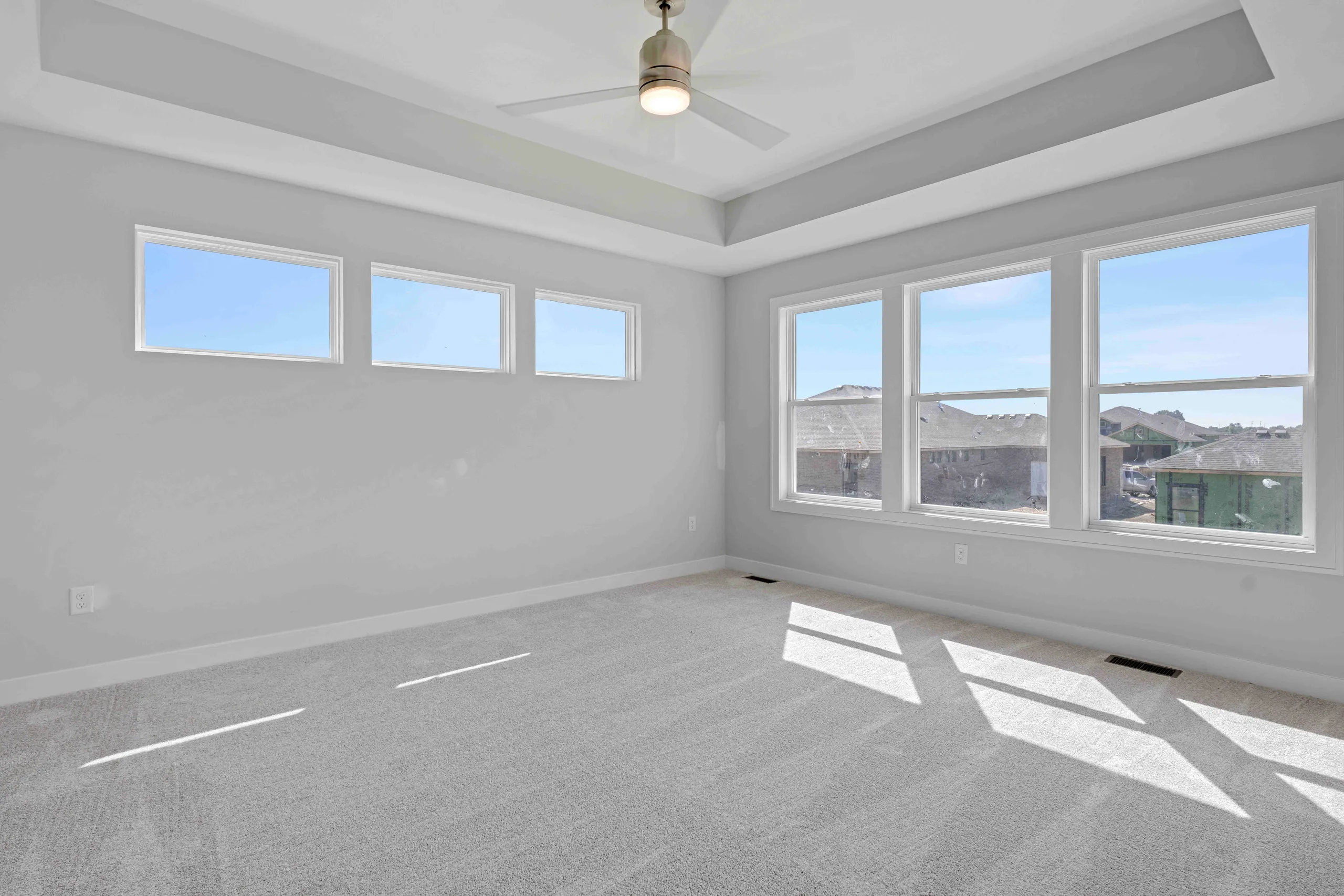 Empty bedroom with gray carpet, white walls, coffered ceiling, ceiling fan, and large windows overlooking neighborhood.