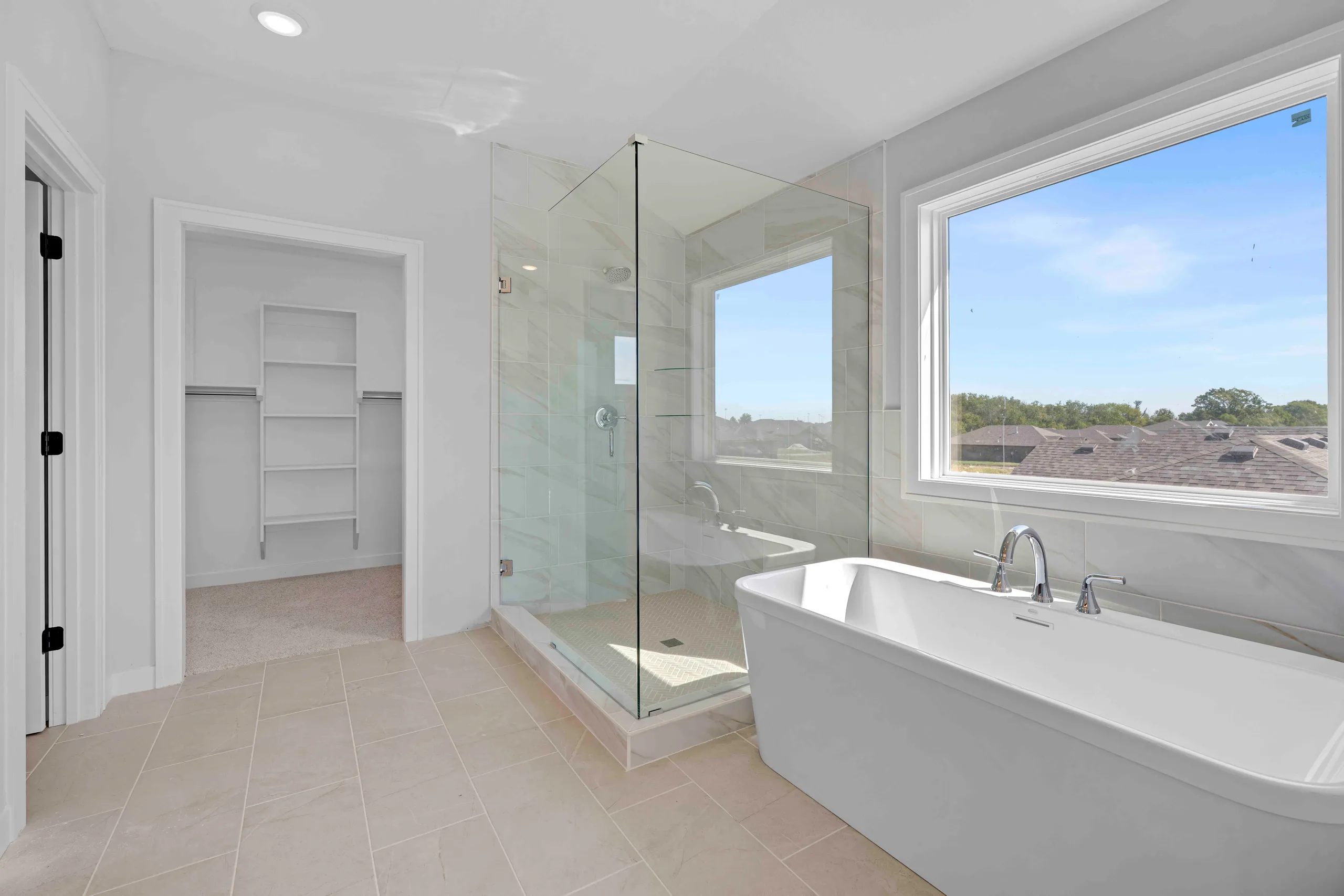 Master bathroom with white freestanding soaking tub, glass walk-in shower, and large window overlooking rooflines.