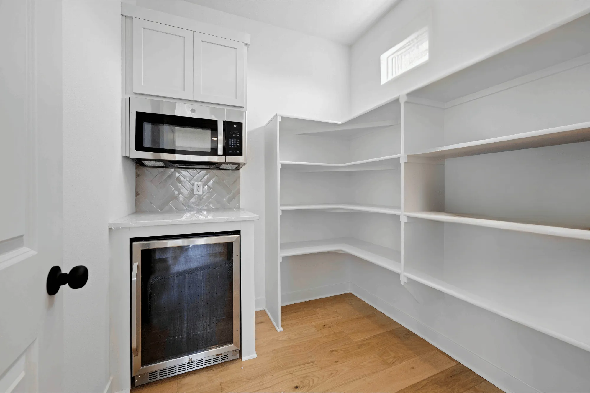 Walk-in pantry with white shelving, built-in microwave and wine cooler, herringbone backsplash, and wood flooring.