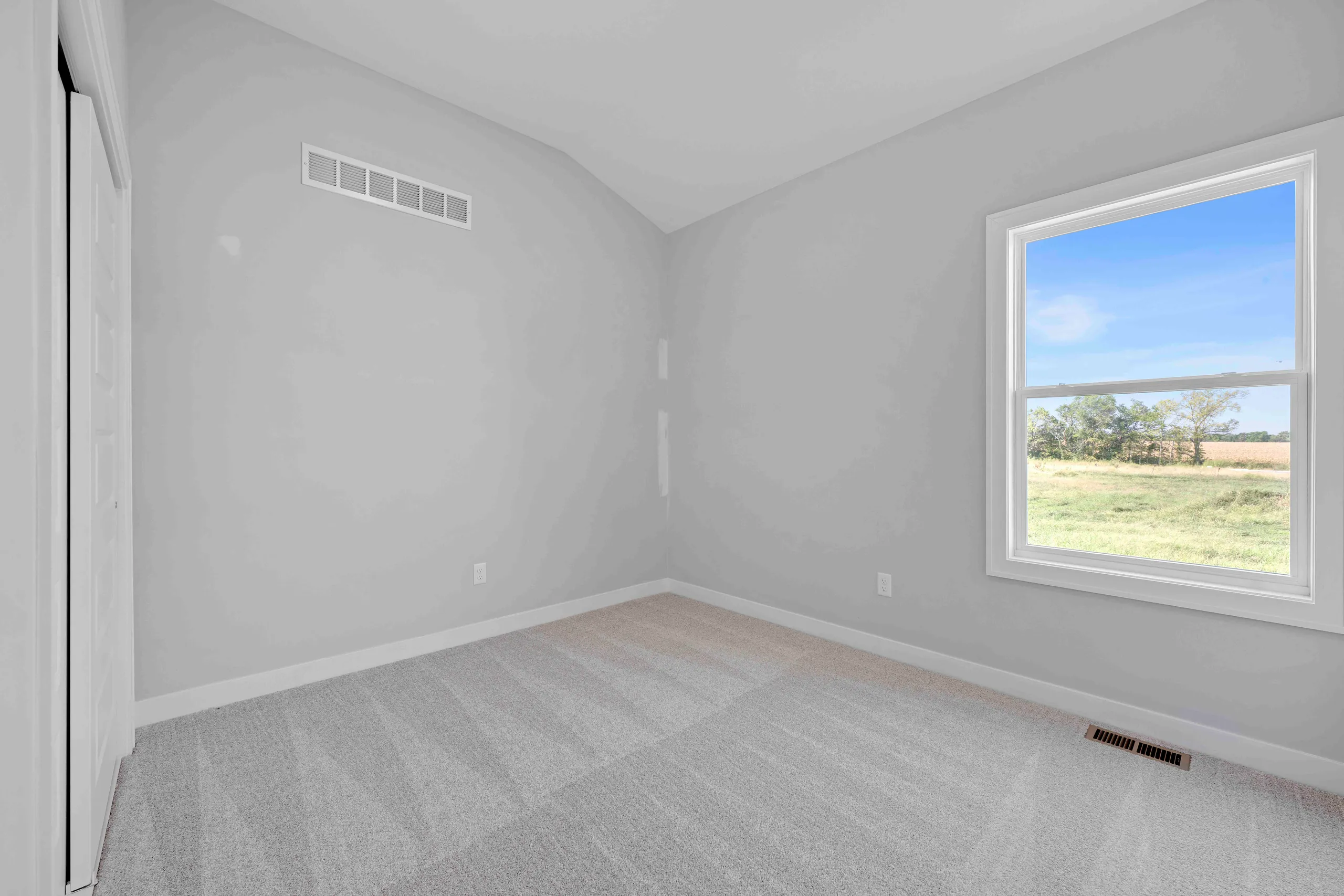 Empty bedroom with gray walls, carpeted floor, and large window overlooking open fields.