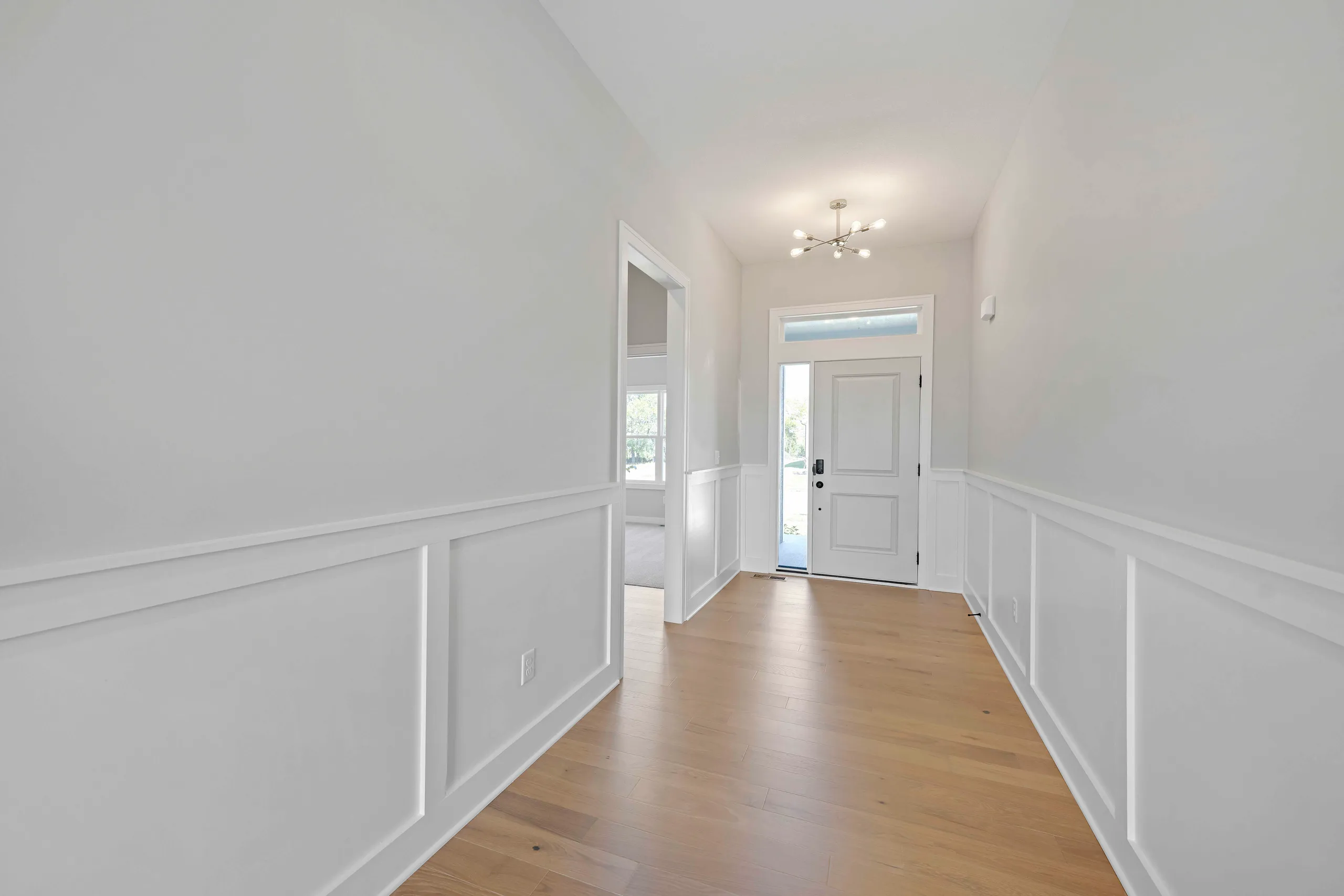 Bright entryway with white wainscoting, wood flooring, and white front door with transom window.