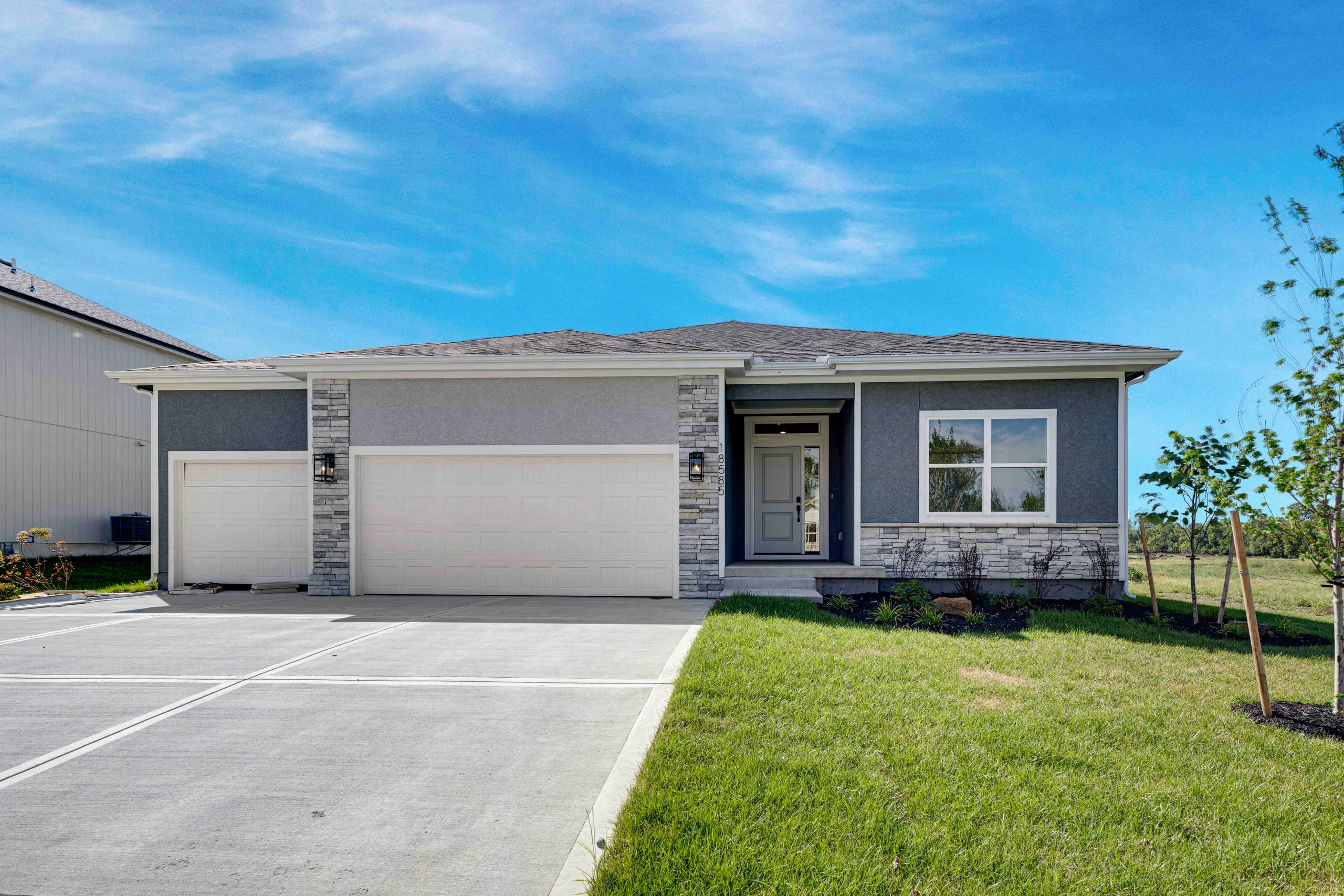 Single-story home with gray siding, stone accents, two-car garage, and landscaped front lawn under clear blue sky.