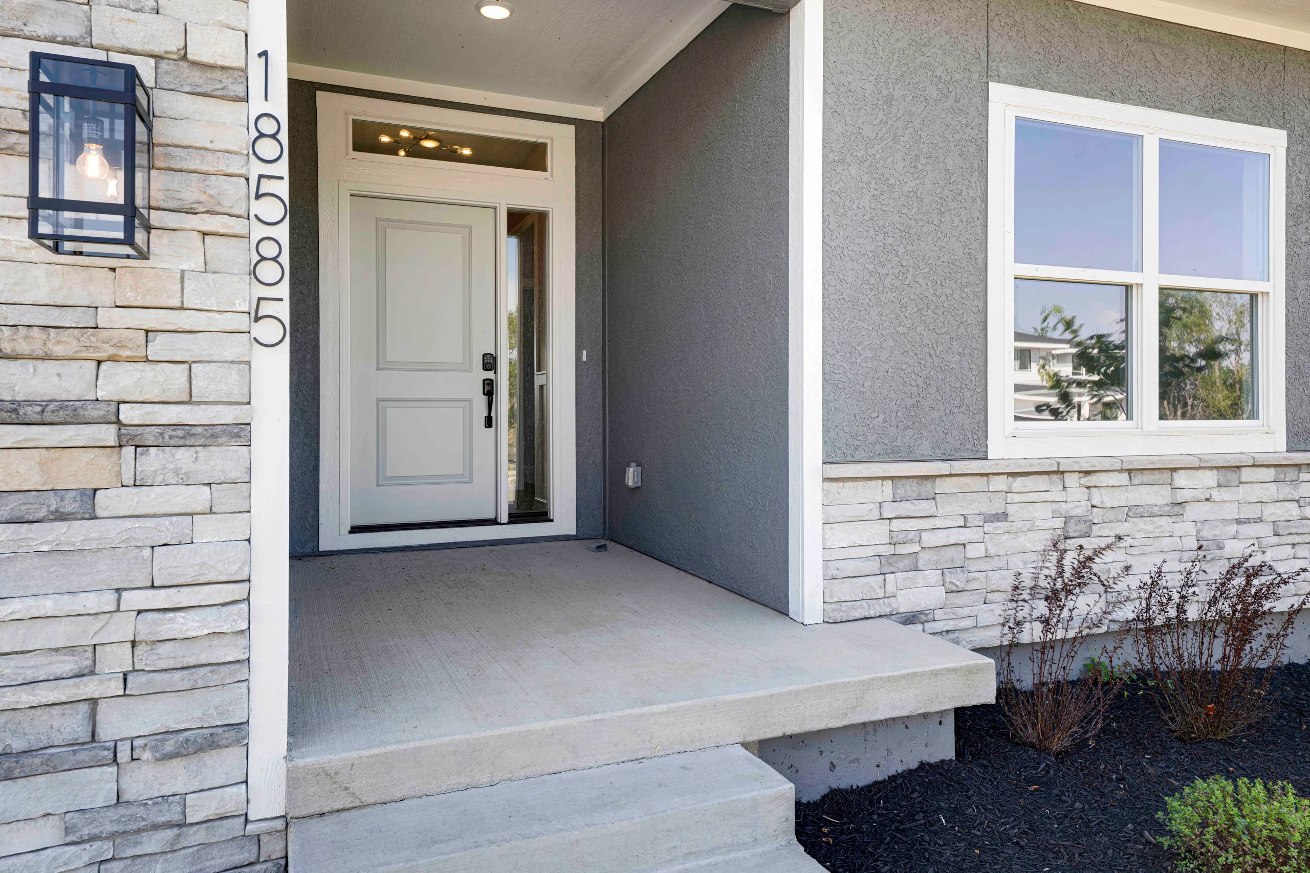Front entry porch with stone and stucco facade, white paneled door, transom light, concrete stoop, and four-pane window.