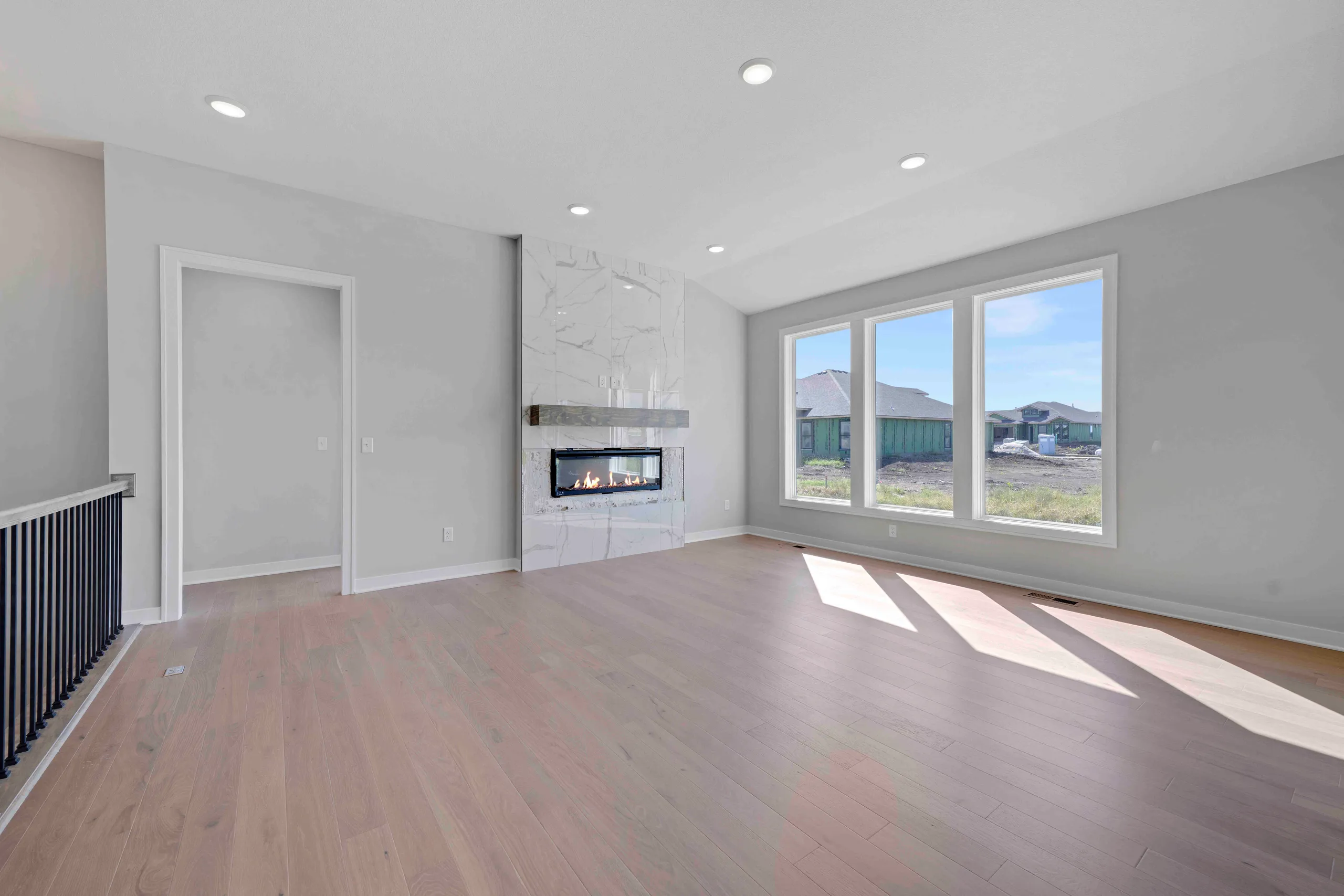 Open living room with fireplace, wood flooring, large windows overlooking community, and white door.