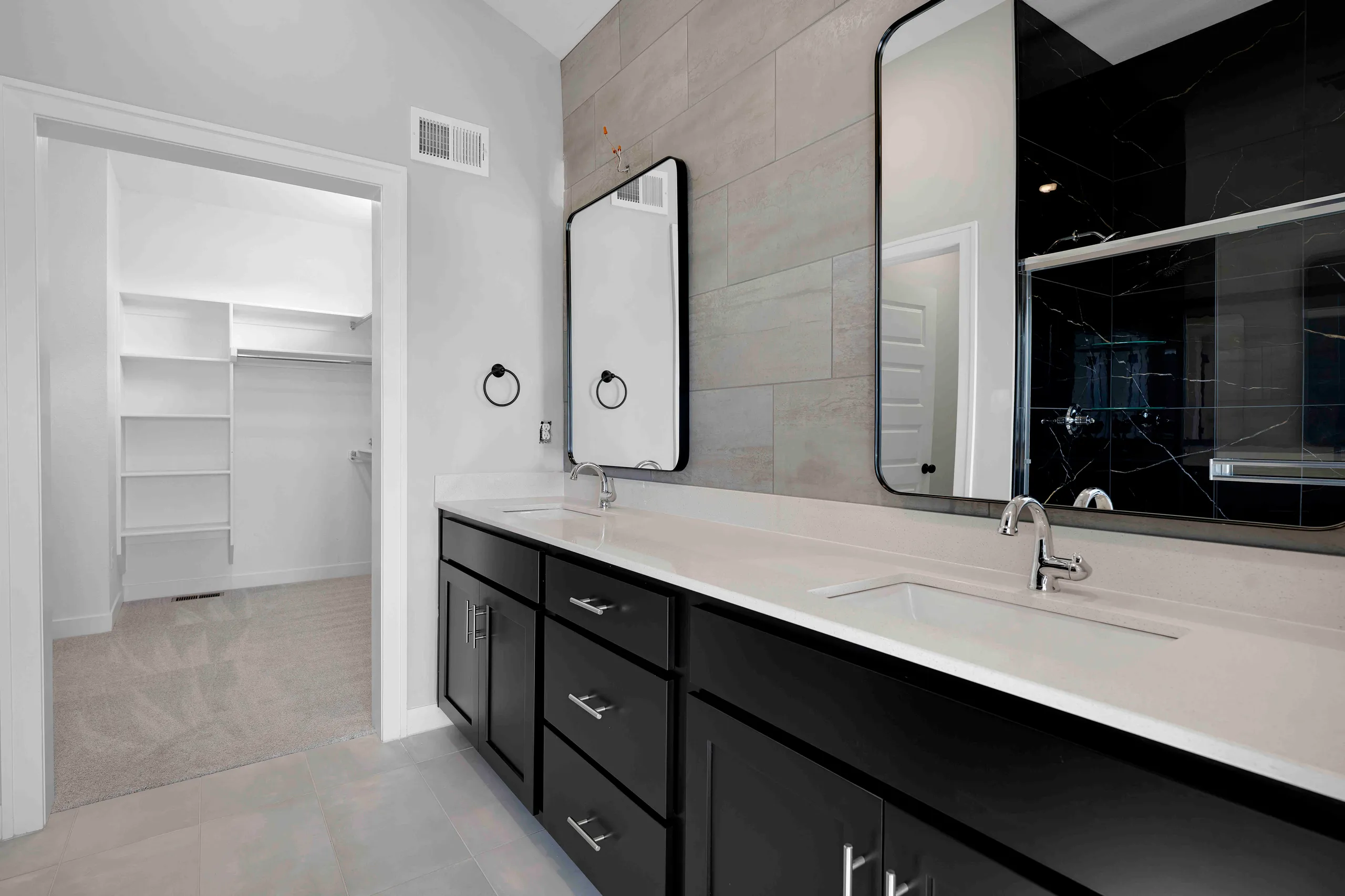 Black vanity with white countertop, dual sinks, and framed mirrors in a bathroom with gray tile walls and walk-in closet.