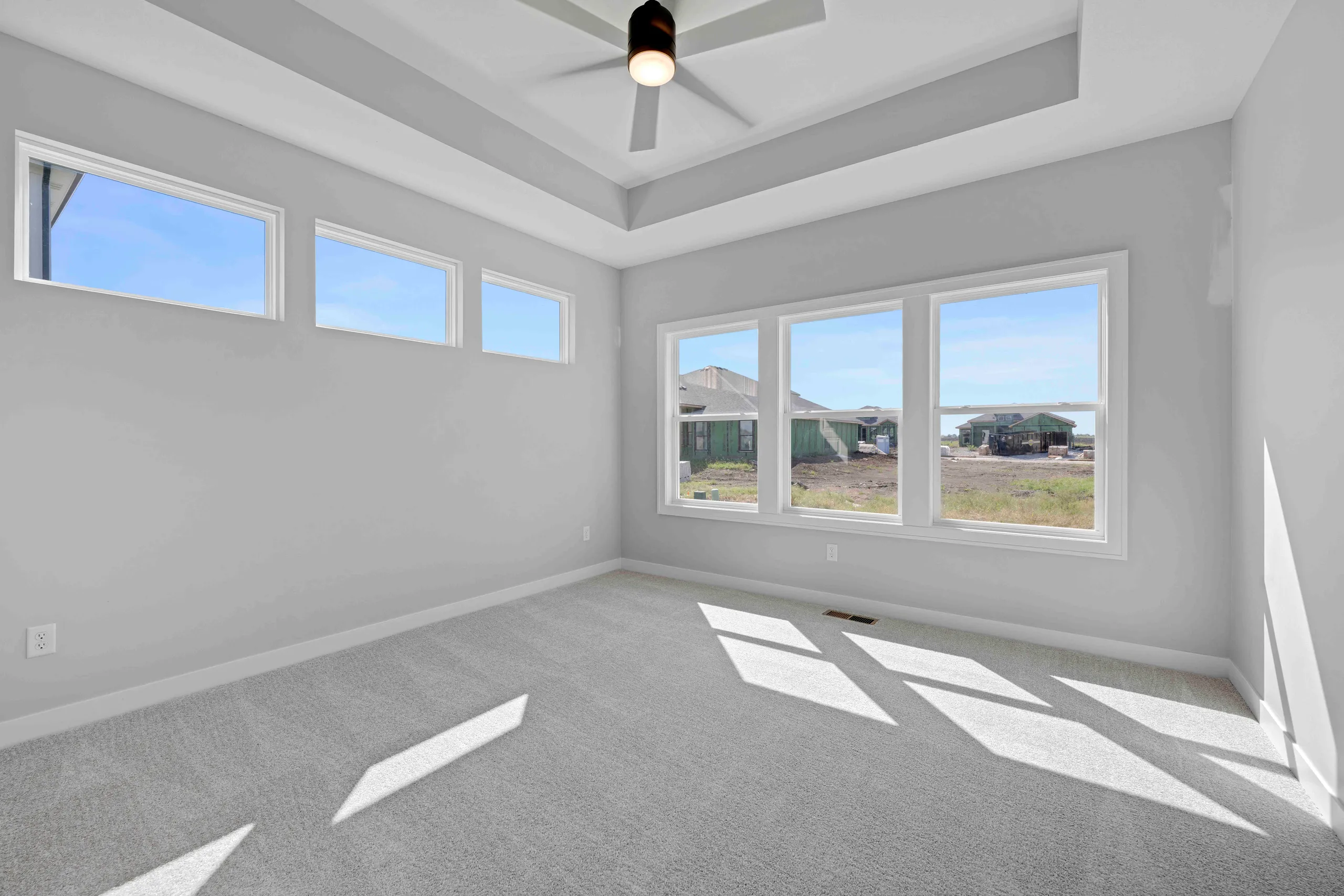 Empty bedroom with gray carpet, coffered ceiling, black ceiling fan, and three windows overlooking neighboring homes.