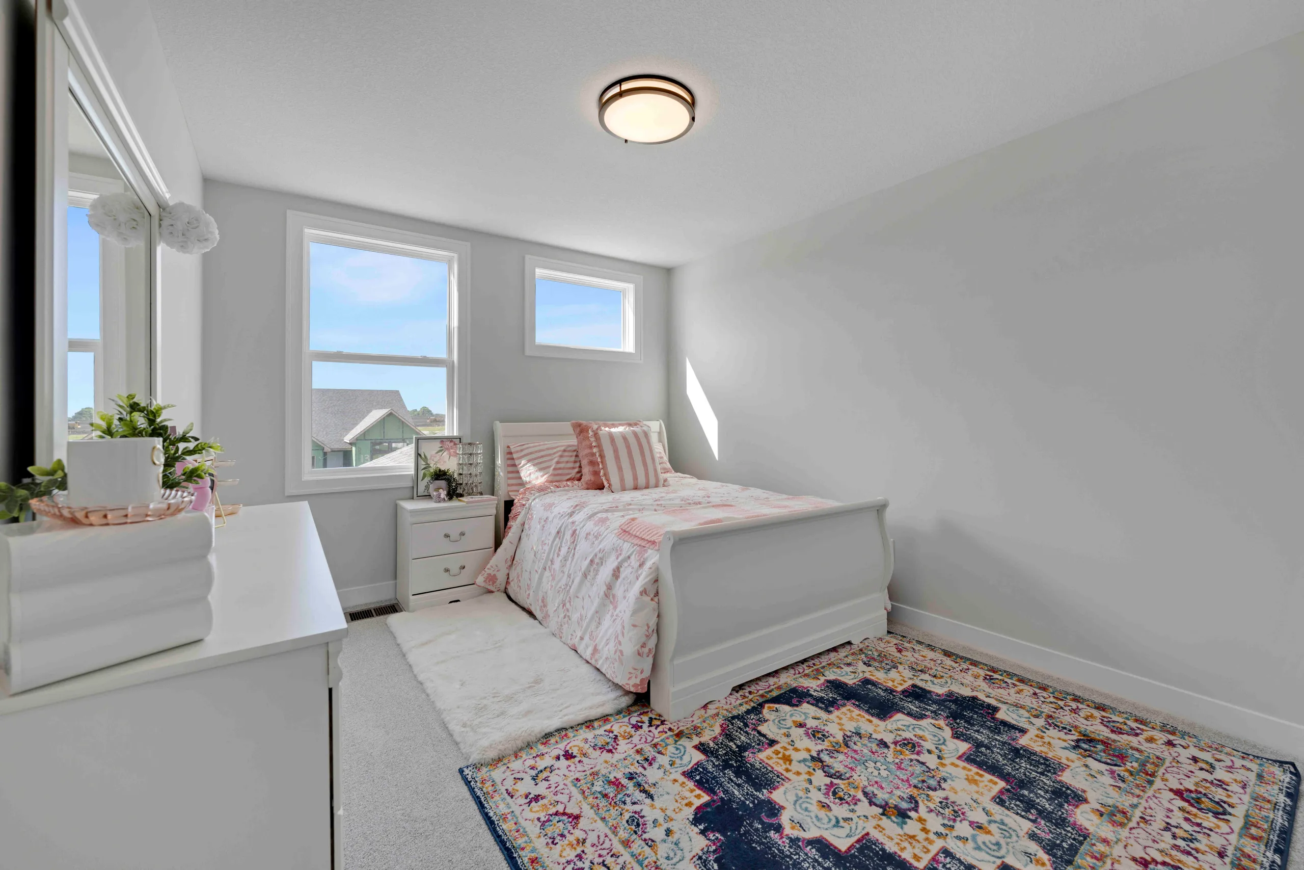 Bright bedroom with white bed, nightstand, potted plants on windowsill, patterned area rug, and two large windows