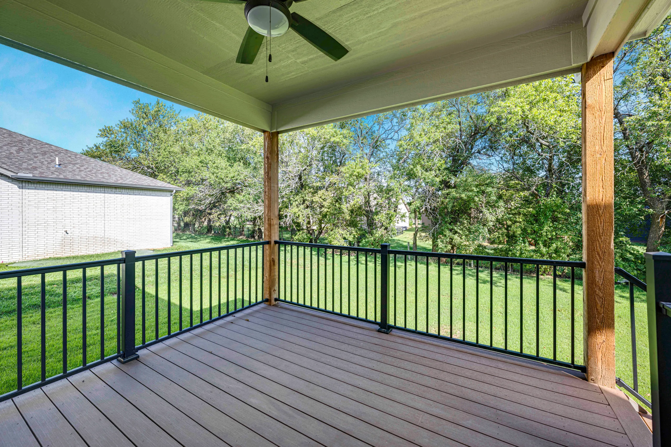 Covered deck with composite flooring, black metal railing, ceiling fan, and view of backyard with trees and garage.