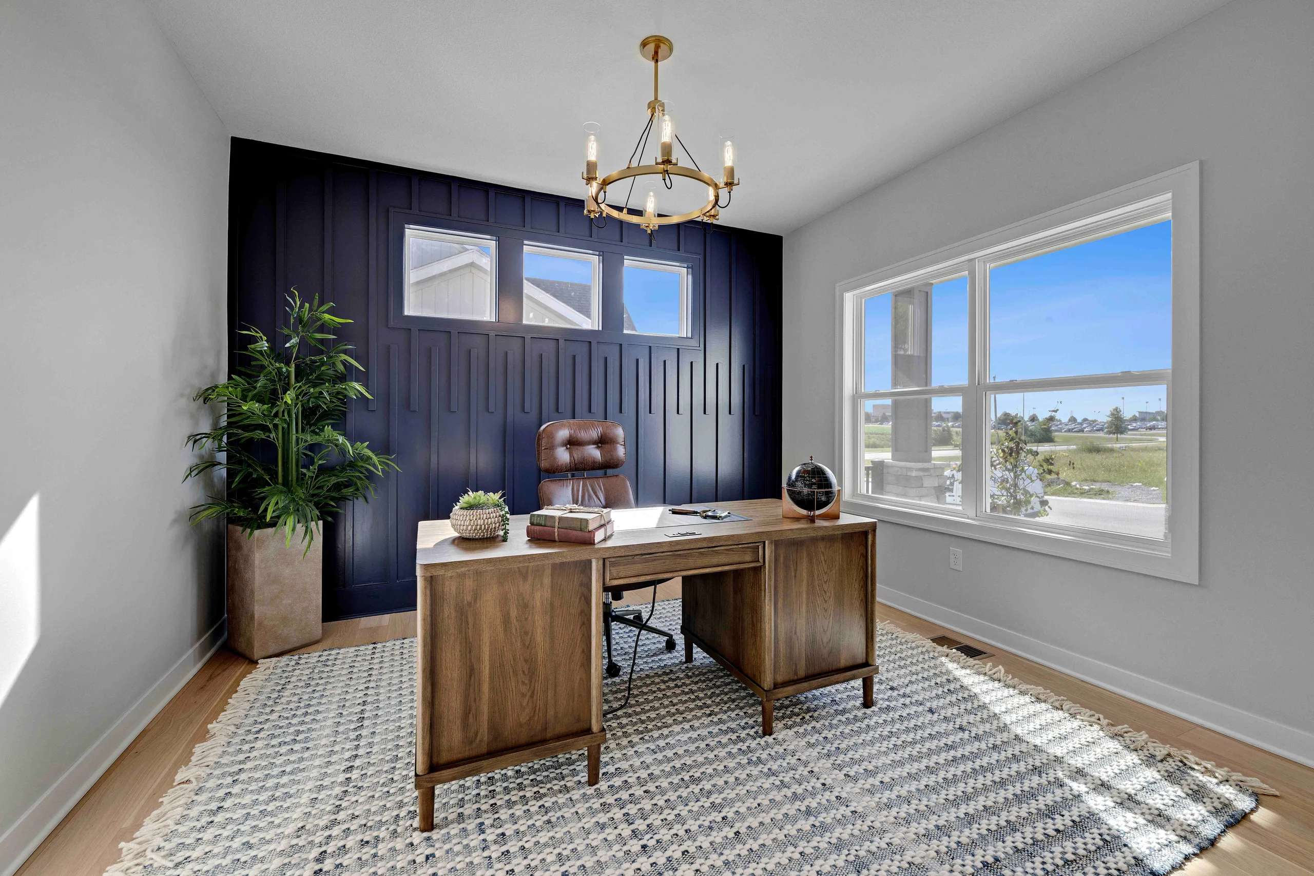 Home office with wood desk, brown leather chair, dark blue shiplap wall, large windows, and brass chandelier.