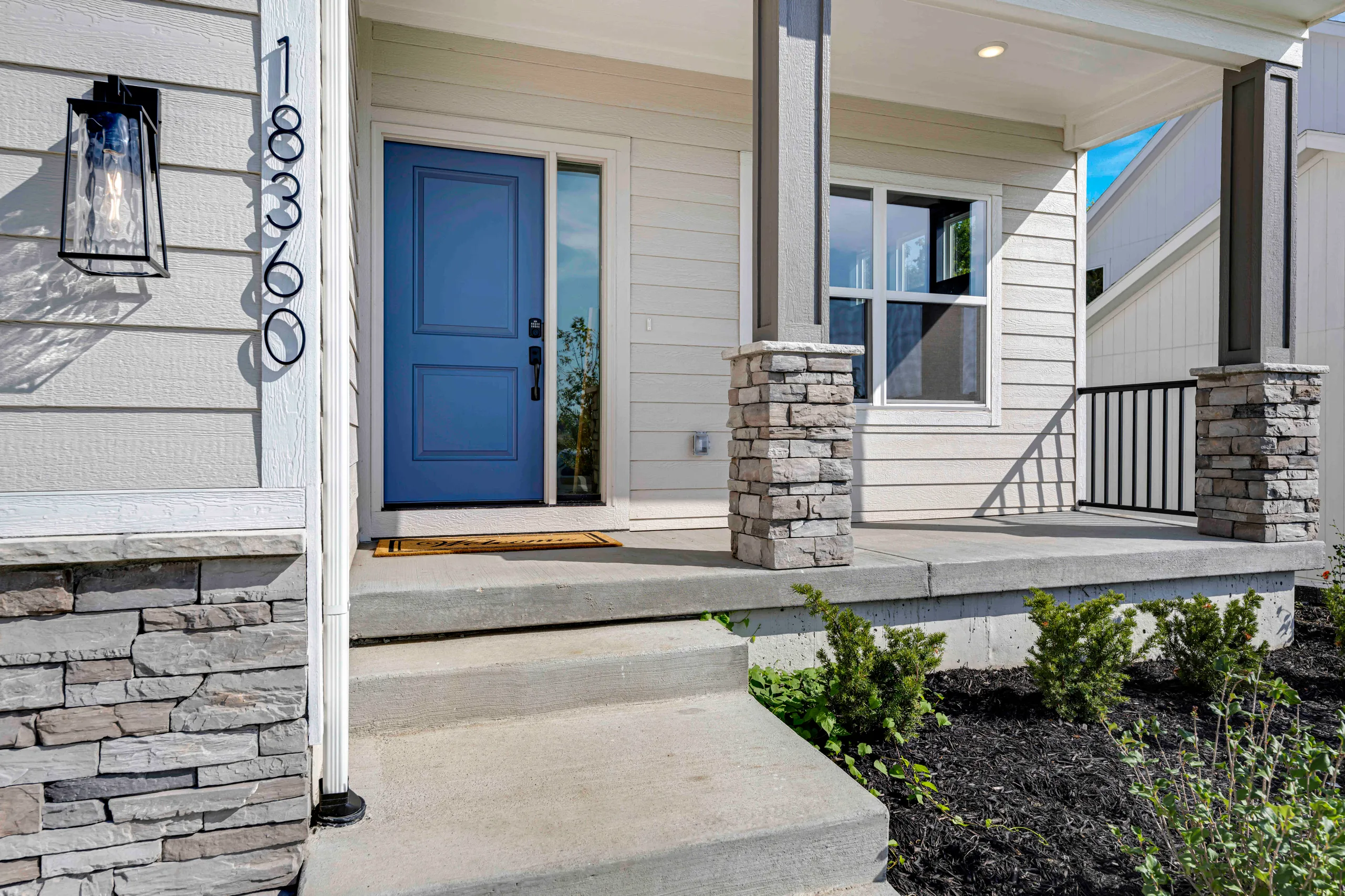 Front entry with blue door, stone pillars, concrete porch, white siding, and black metal railing.
