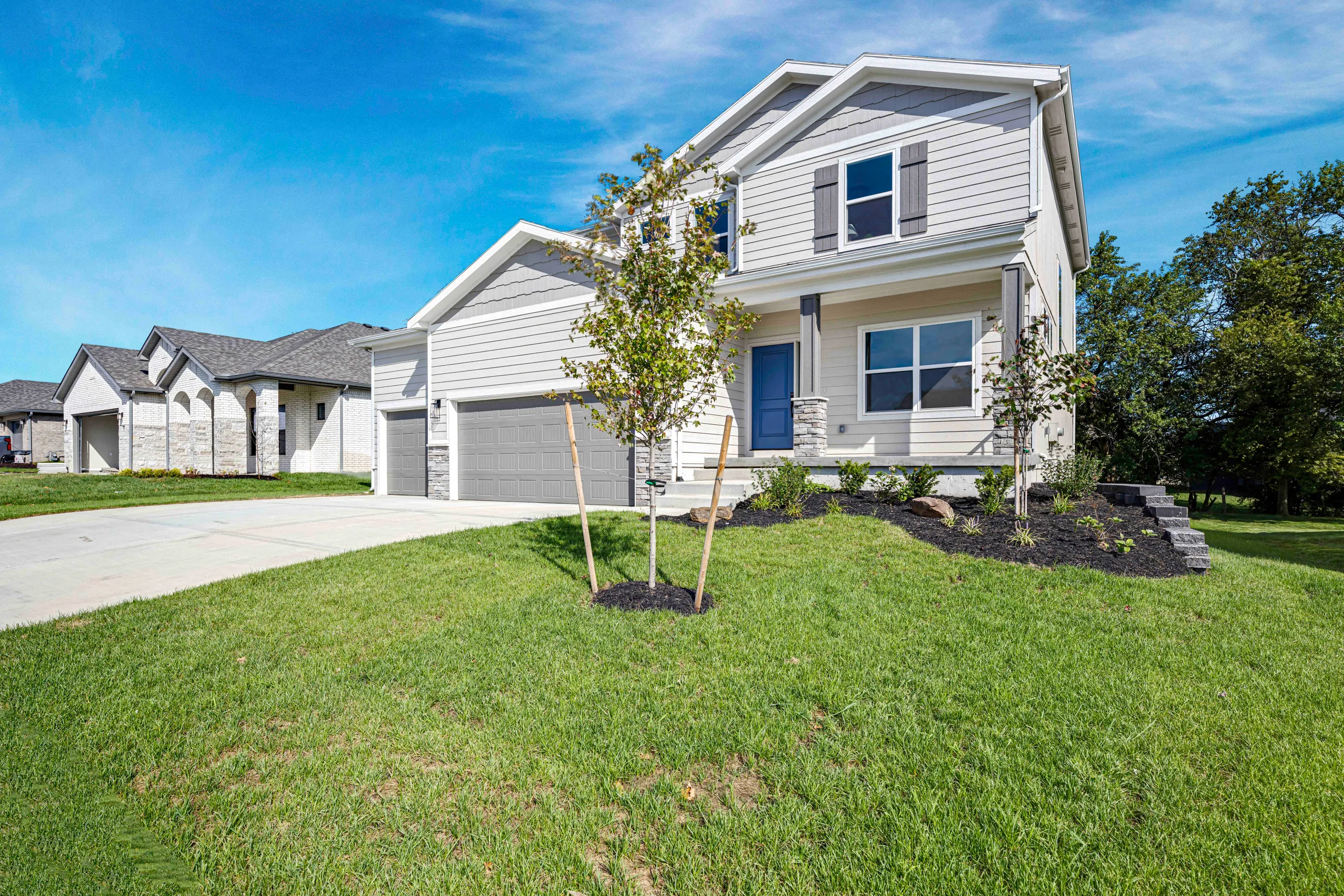Two-story white house with blue front door, two-car garage, and young trees in landscaped front yard.