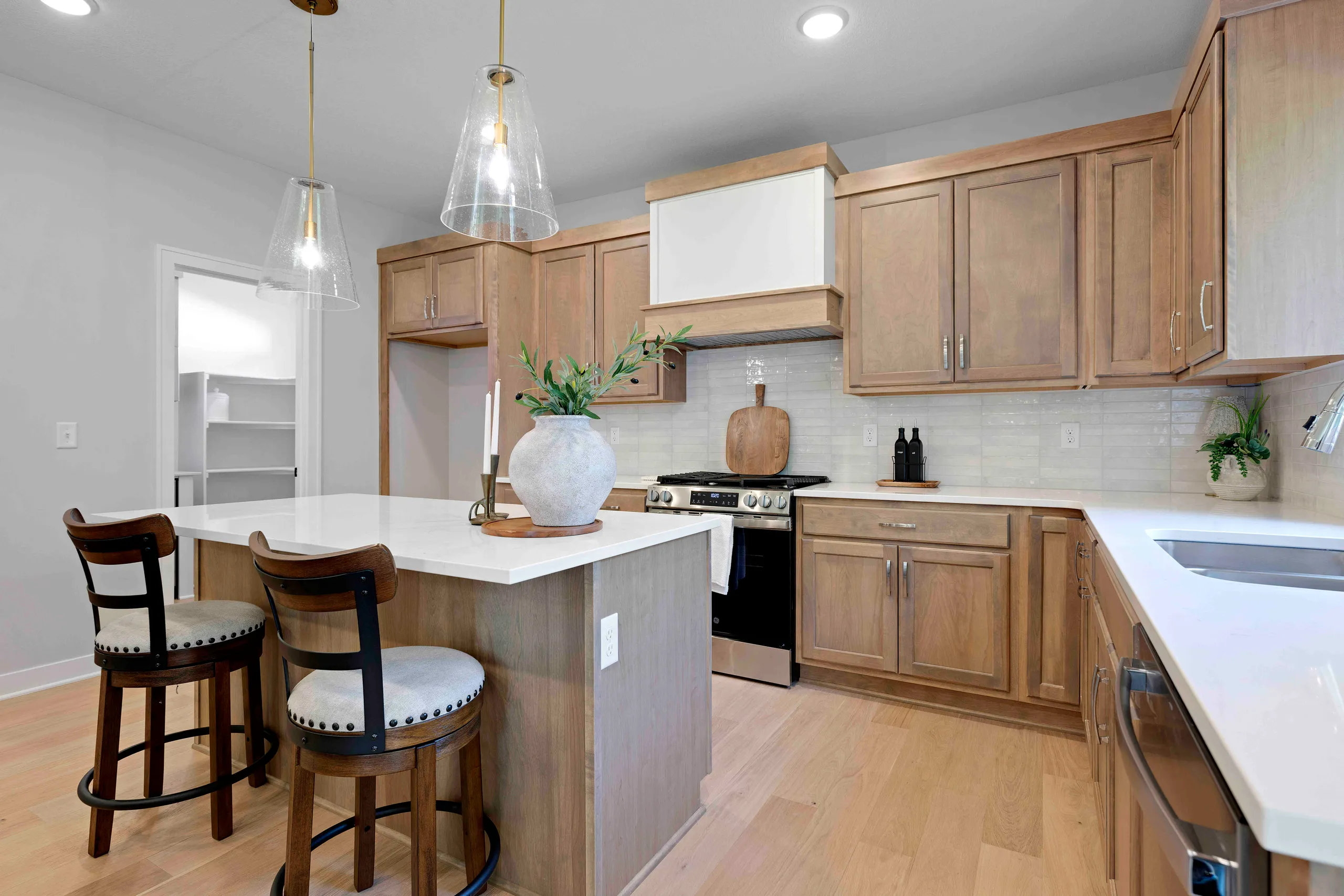 Kitchen with wood cabinetry, white quartz island with two wood stools, stainless steel appliances, and pendant lighting.