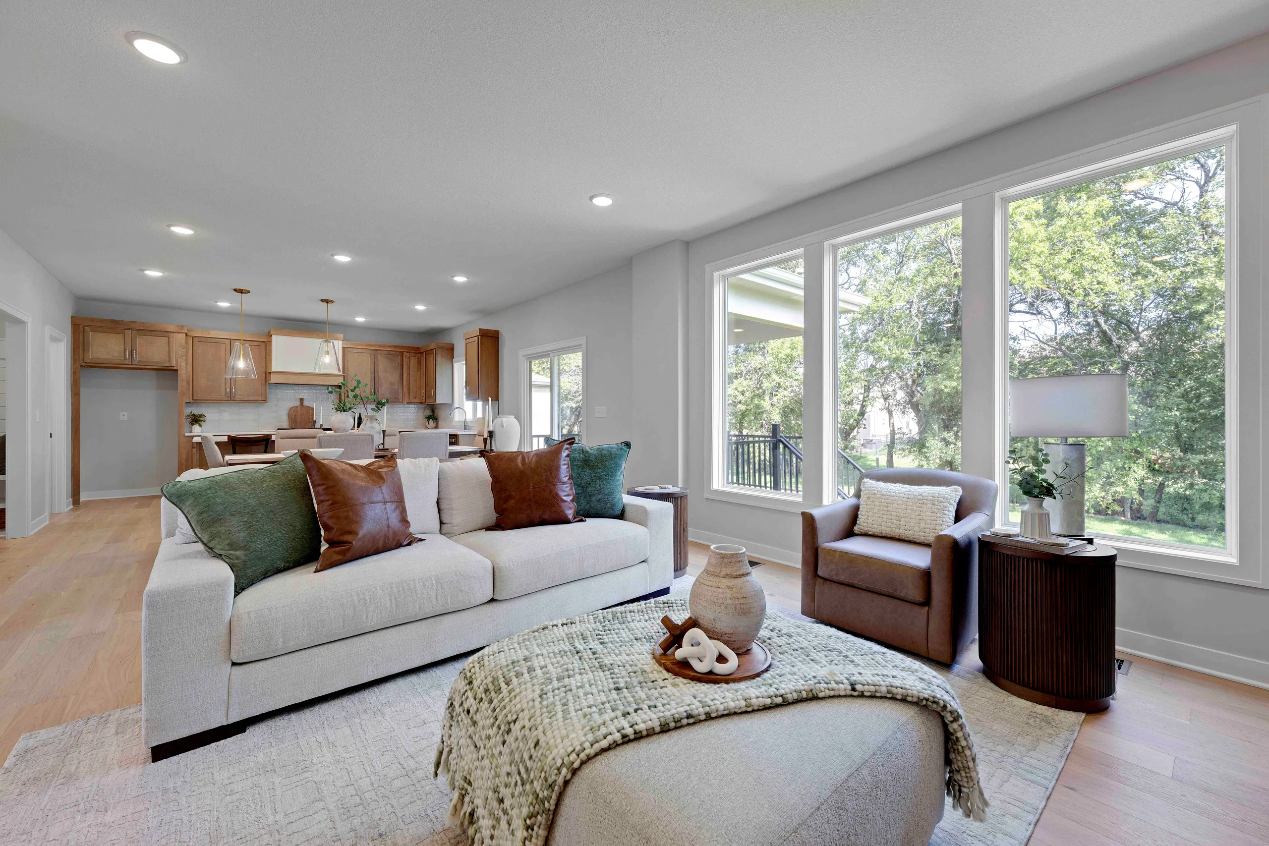 Open-concept living room with cream sofa, brown leather chair, and large windows overlooking trees, adjacent to kitchen with