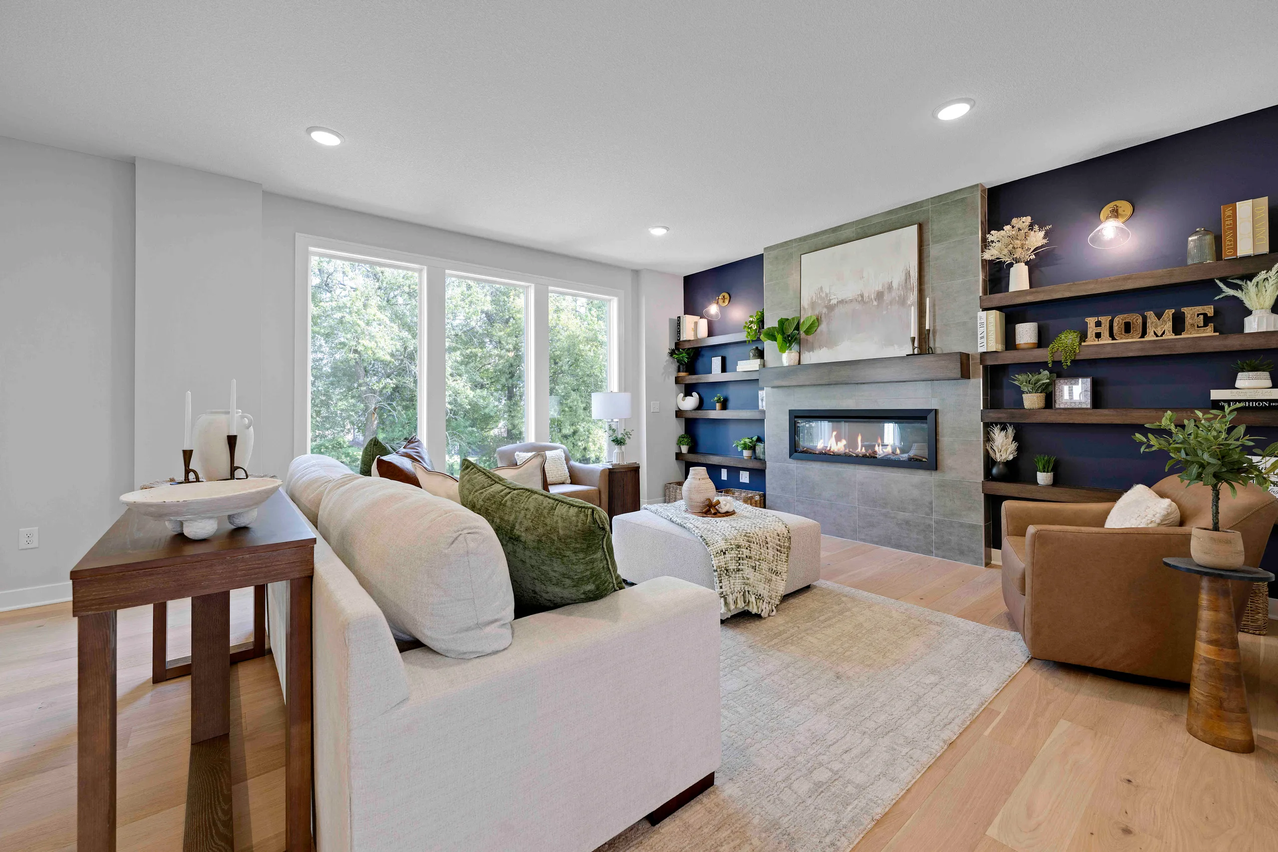 Living room with cream sofa, leather armchair, fireplace with blue accent wall, wood shelving, and large windows overlooking