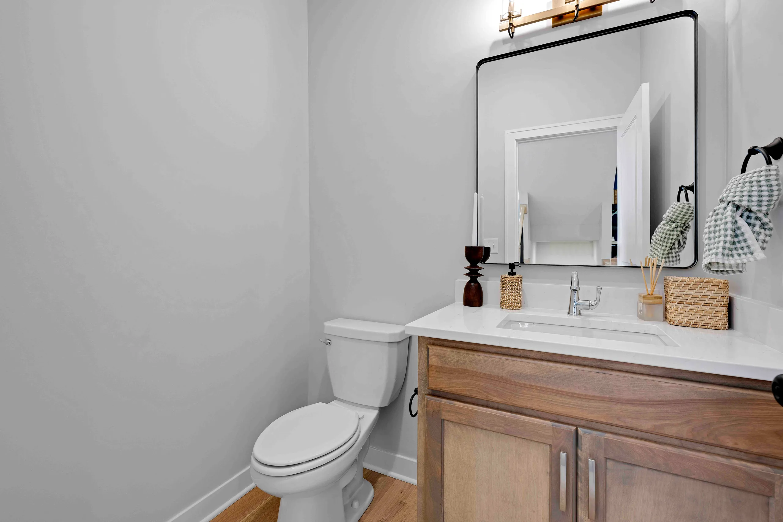 Bathroom with white walls, wood vanity, white countertop sink, black-framed mirror, and toilet on wood flooring.