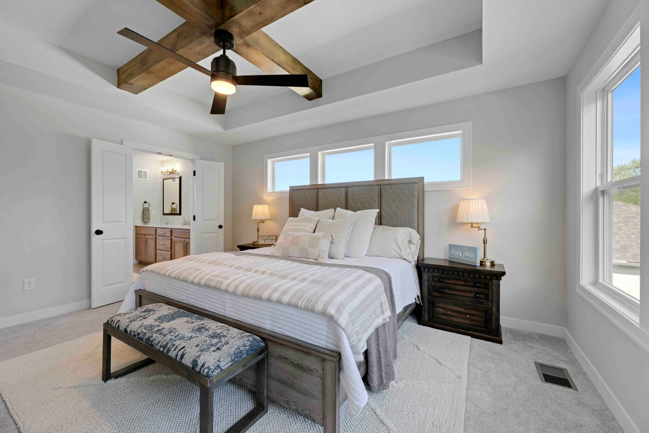 Master bedroom with gray upholstered bed, wood beam ceiling, ceiling fan, nightstands with lamps, and large windows.