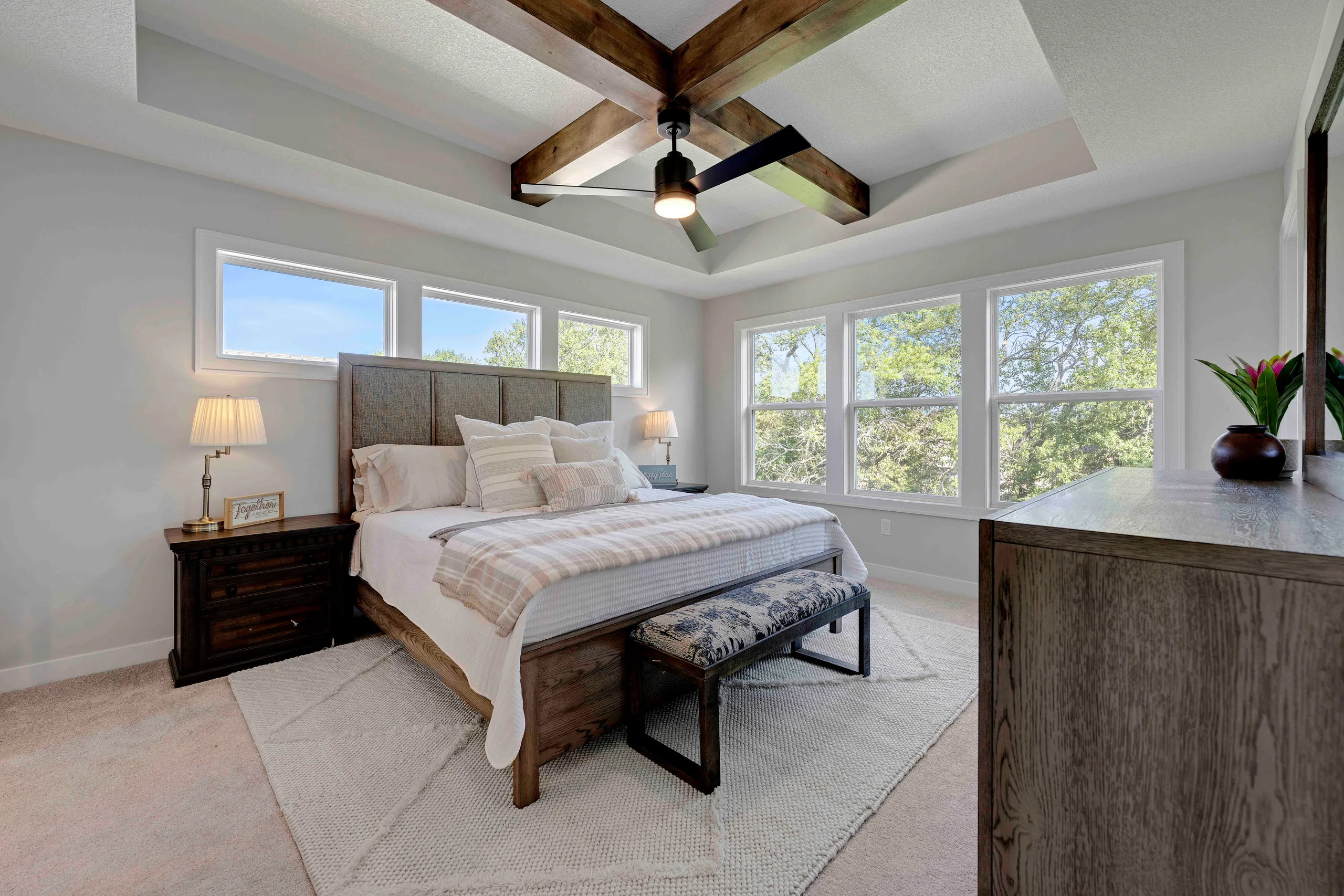 Master bedroom with wood beam ceiling, gray upholstered bed, nightstands, ceiling fan, and large windows overlooking trees.