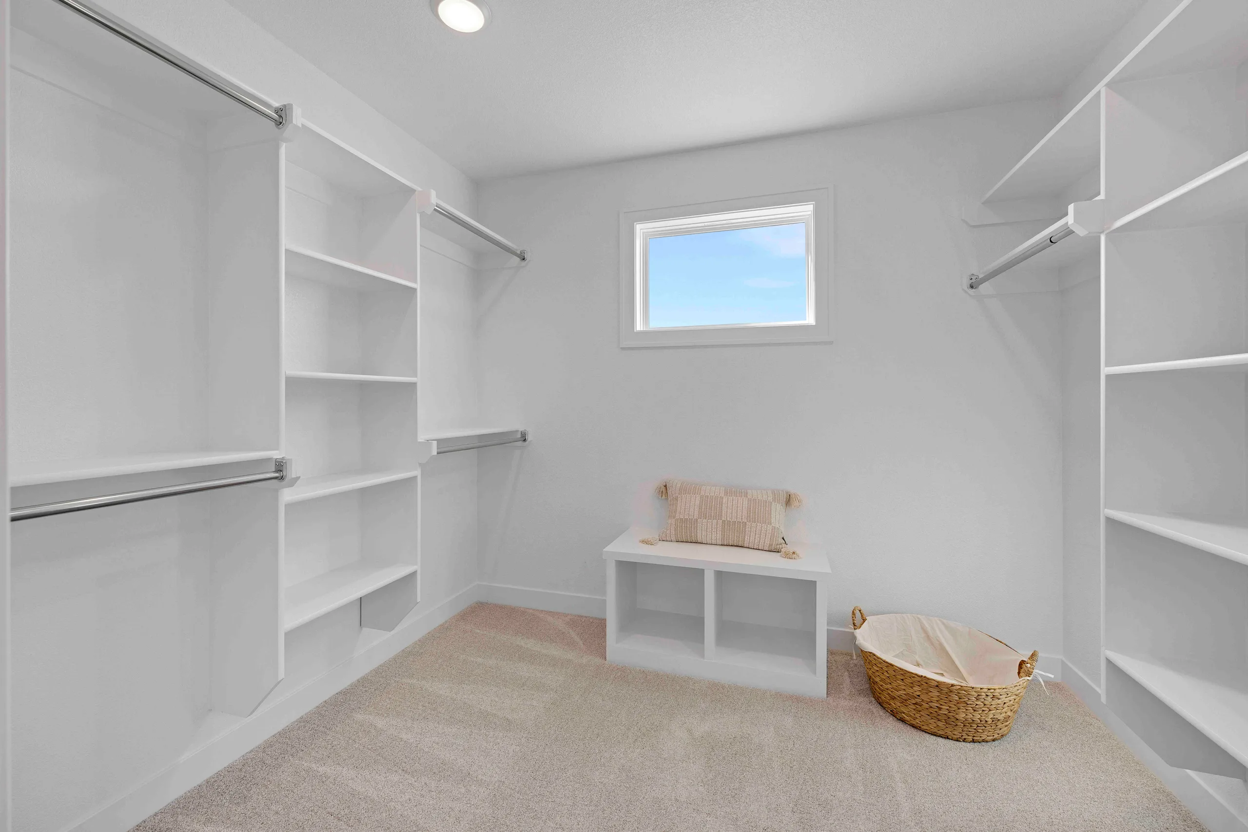 Walk-in closet with white shelving and hanging rods on both sides, window, bench with cushion, and woven basket.