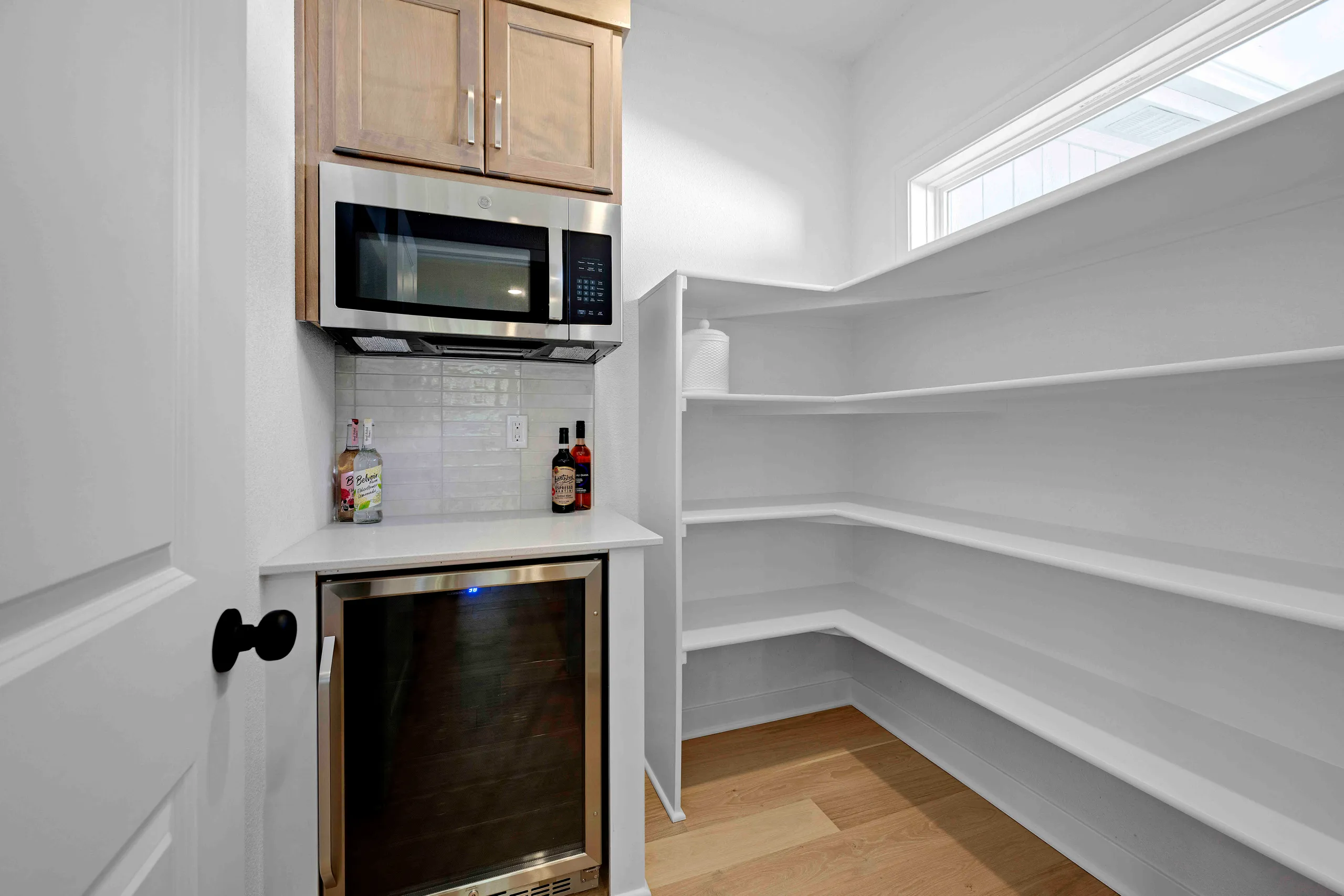 Pantry with white shelving, wood flooring, microwave, wooden cabinets, and beverage cooler.