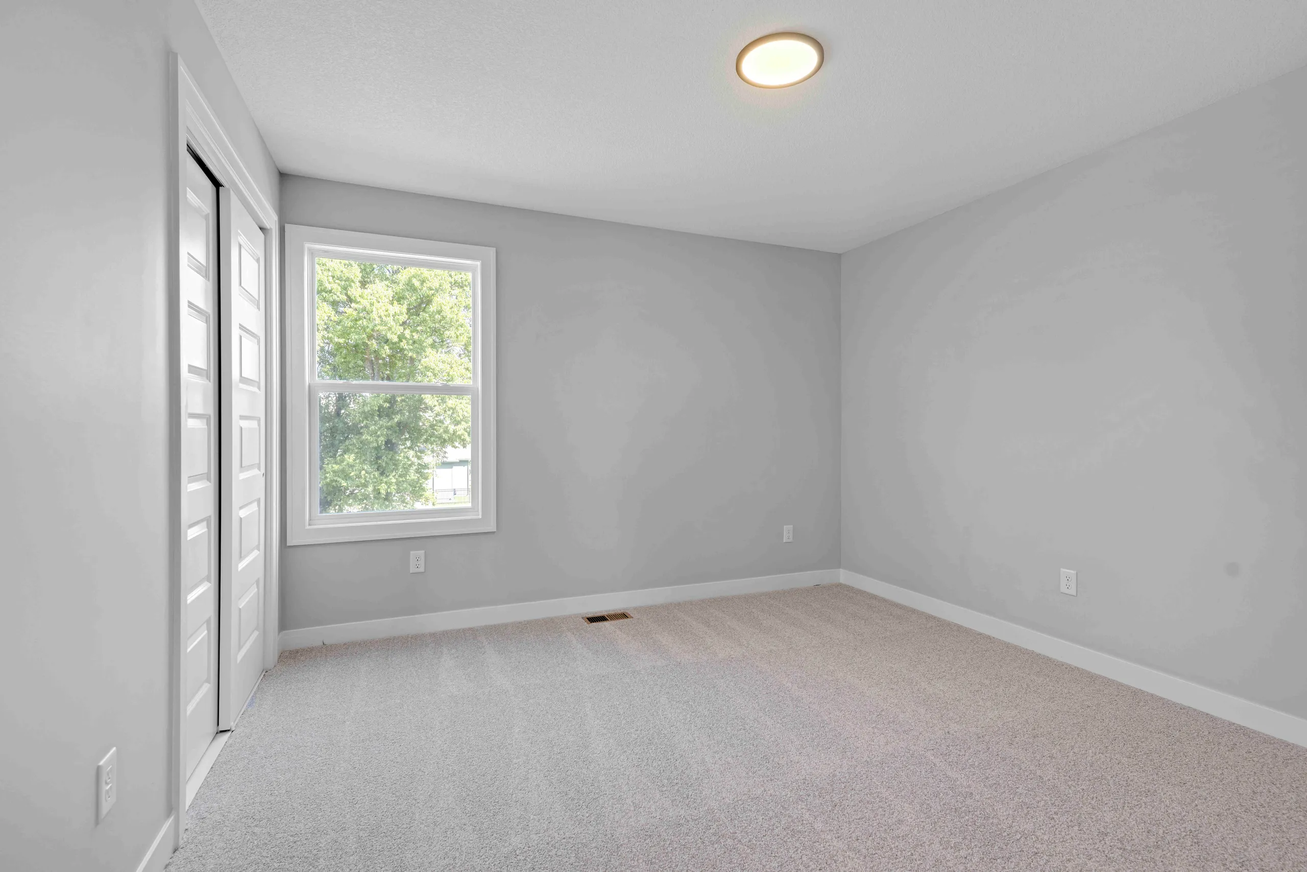 Empty bedroom with gray walls, carpeted floor, window with white trim, closet doors, and recessed ceiling light.