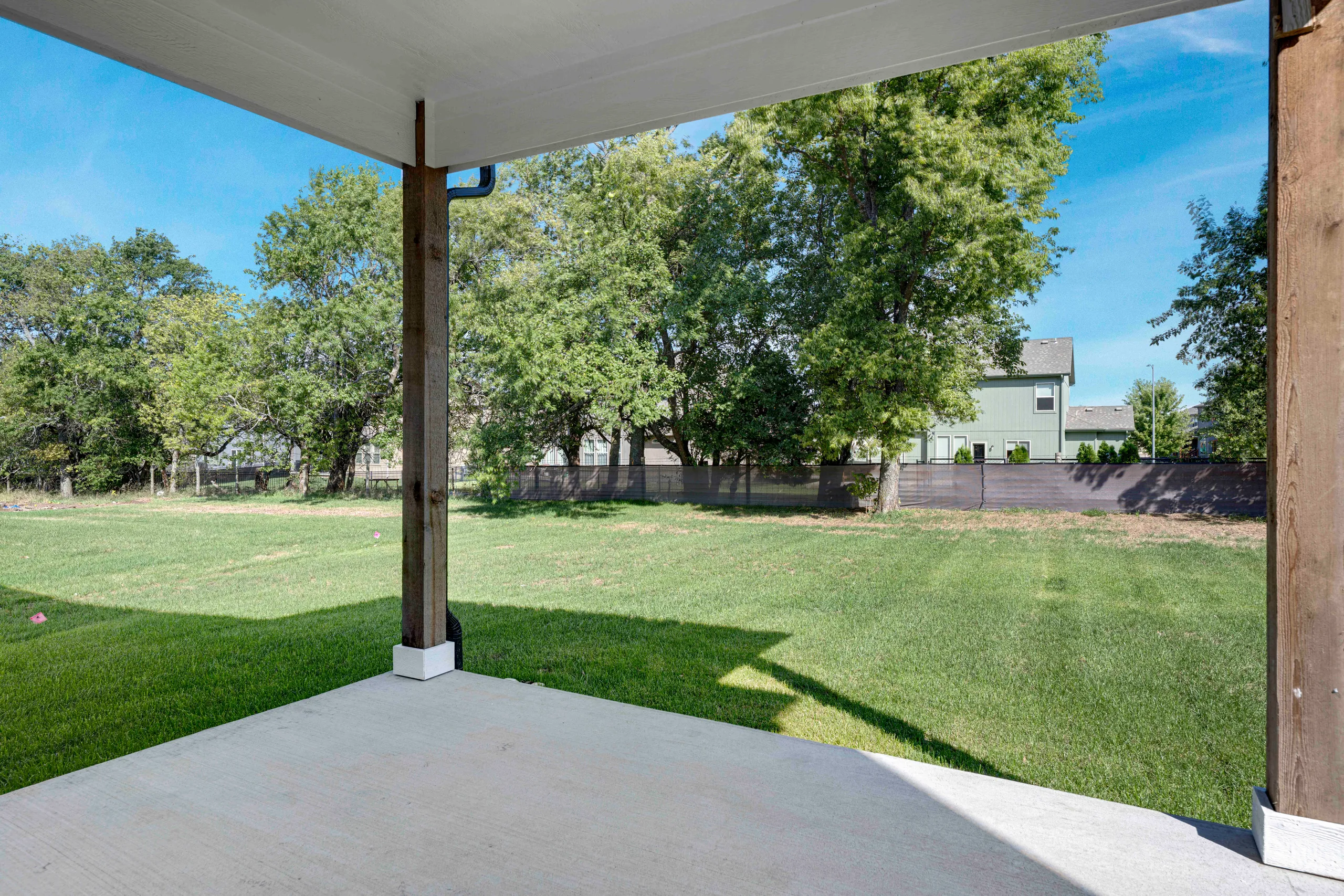 Covered patio with concrete floor overlooking a large backyard with mature trees and a house in the distance.