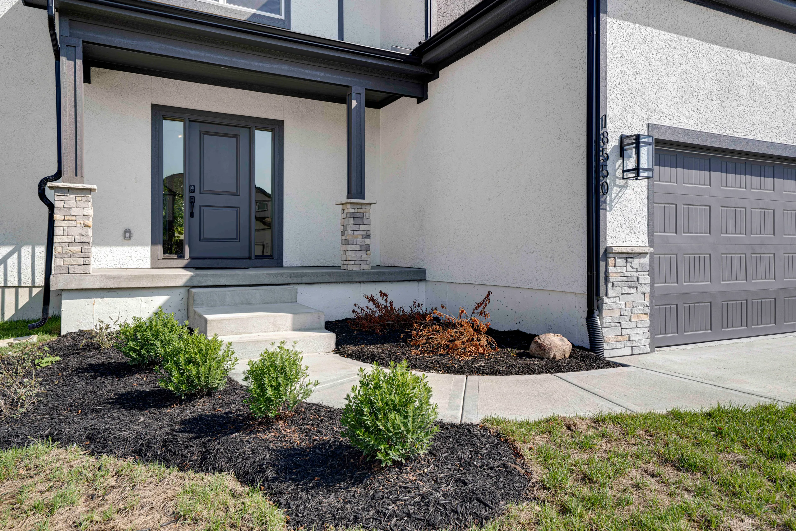 Home exterior with gray stucco walls, dark front door, stone pillars, attached garage, and landscaped front yard with mulch