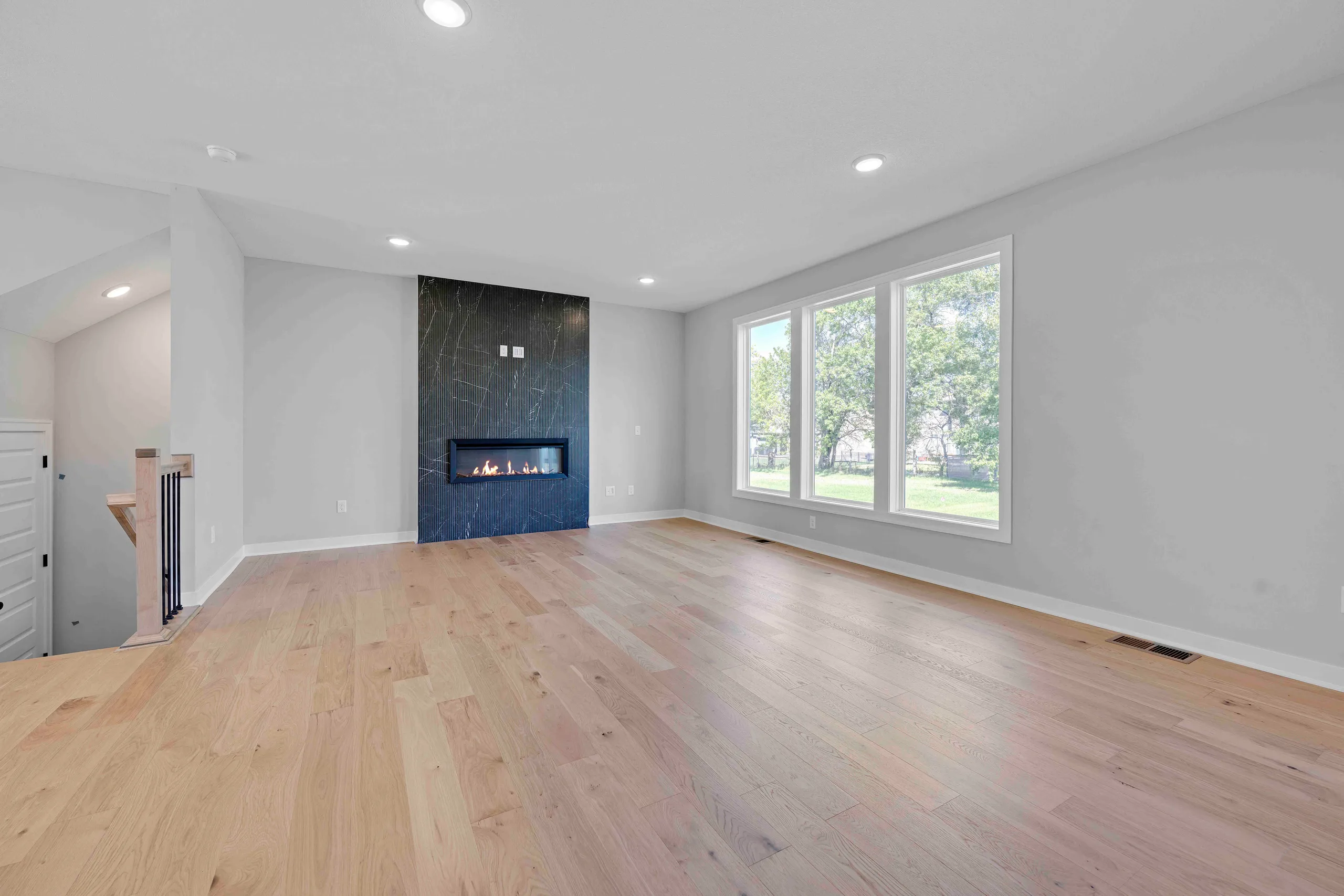 Open living room with hardwood floors, fireplace with dark tile surround, and large multi-pane windows overlooking trees.