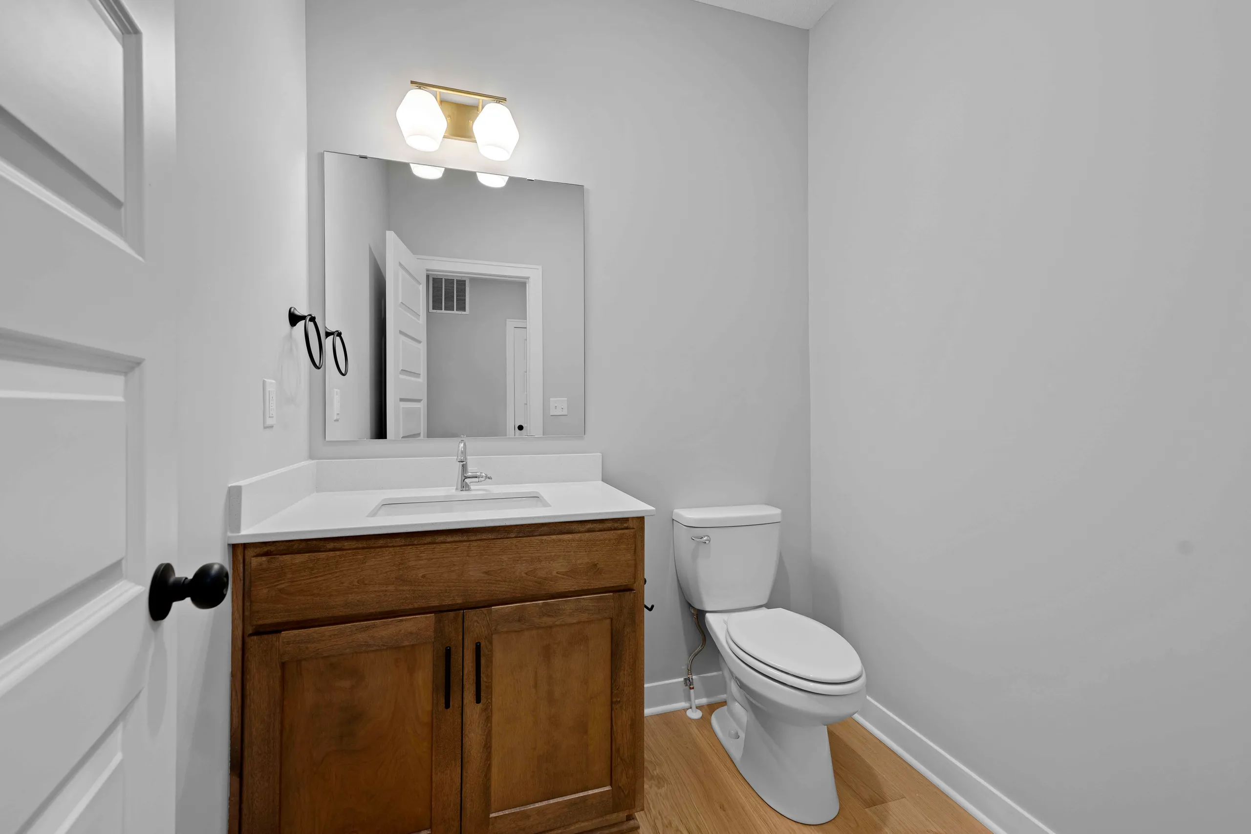 Bathroom with wood vanity cabinet, white countertop, framed mirror, brass light fixture, and toilet on wood plank flooring.