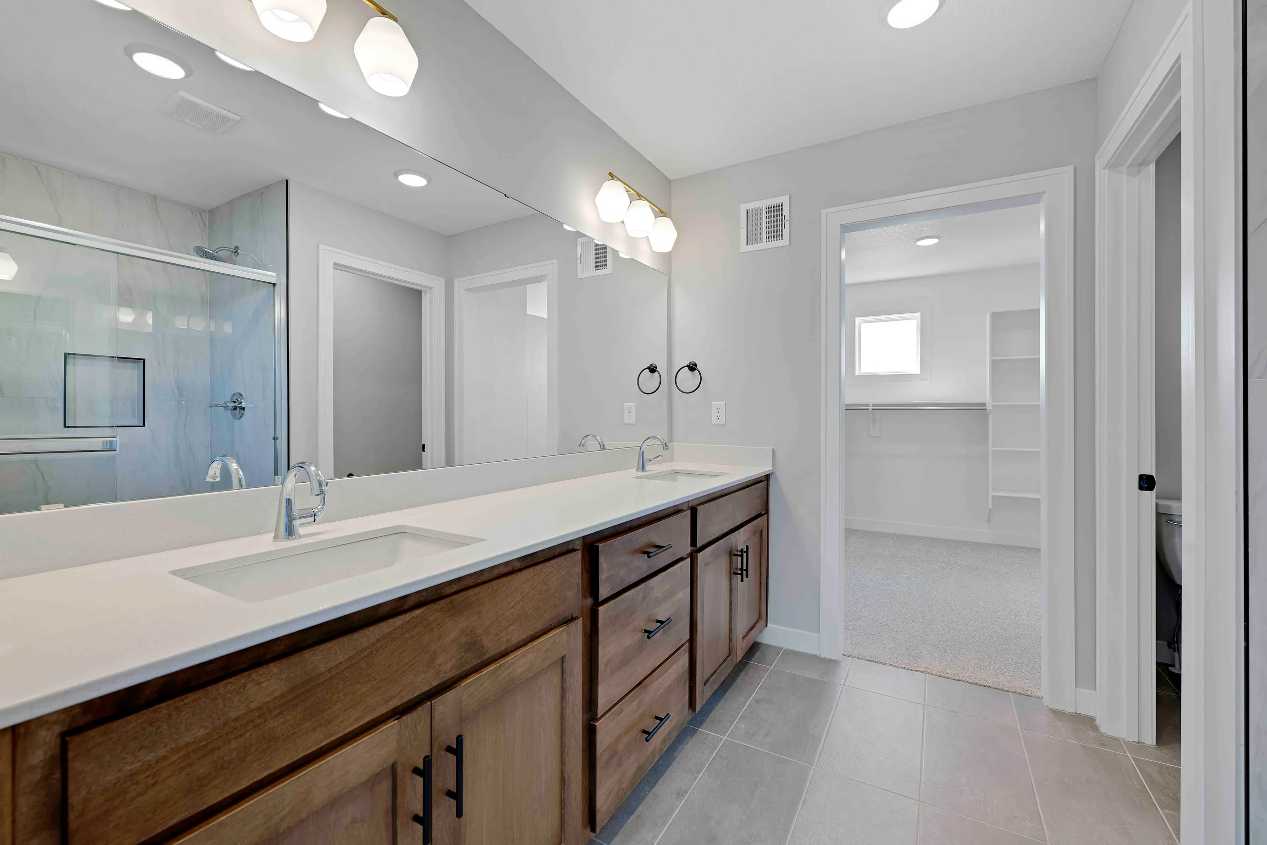 Master bathroom with dual vanity, white countertop, wood cabinetry, large mirror, walk-in closet, and tile flooring.