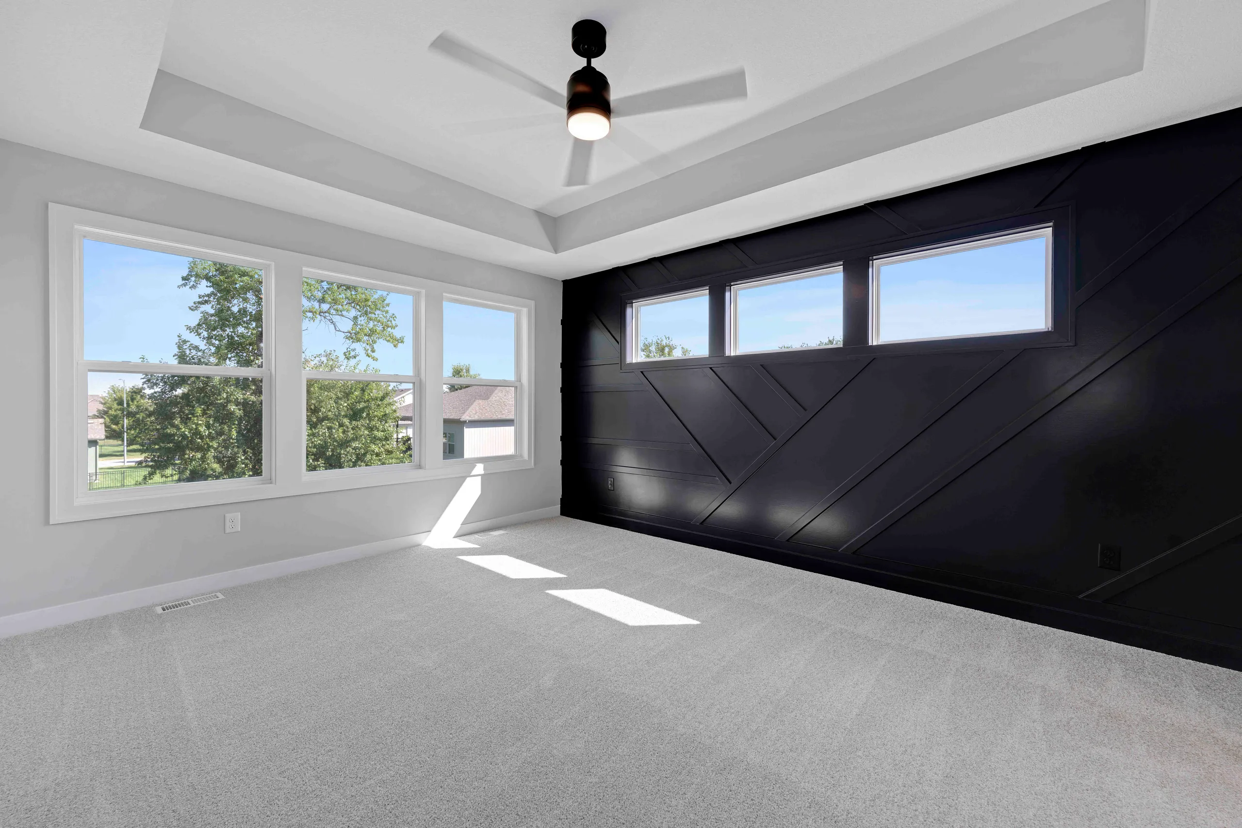 Empty bedroom with gray carpet, white walls, black accent wall with shiplap, large windows, and ceiling fan.