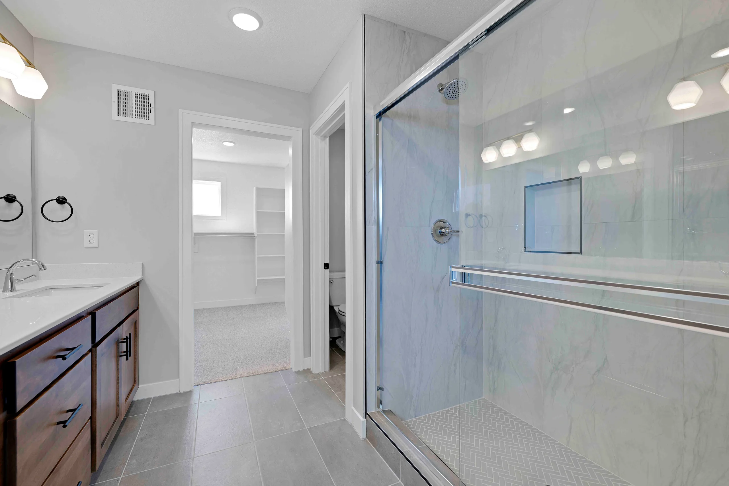 Master bathroom with dark wood vanity, white sink, glass-enclosed shower, and walk-in closet visible through doorway.